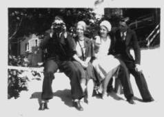 A man sits on a bench with three friends. He stares out at you with a pair of binoculars.