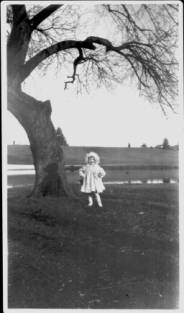 A little girl stands under a tree in her white sunday dress and hat. She has her hands on her hips and seems to be posing with a preternatural self-satisfaction.