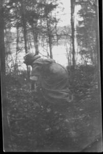 A woman in Victorian dress crawls through the leaves under a barbed wire fence. She is in a scrub-forest. There is a lake in the background. Her face is obscured by shadows. No skin shows. She is hiding, escaping, vanishing into the forest.